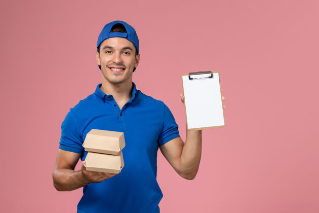 快乐正面图：身穿蓝色制服披风的年轻男性快递员拿着小的快递食品包 在浅粉色的墙上微笑着写着记事本人男性前面