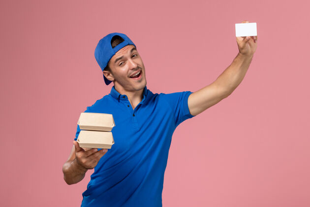 年轻正面图：身穿蓝色制服披肩的年轻男性快递员拿着小包裹 浅粉色墙上有卡片正面人持有