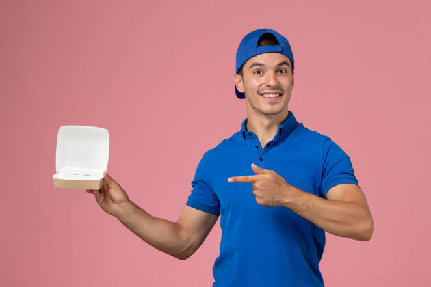 男人正面图身穿蓝色制服披肩的年轻男性快递员拿着粉色墙上的小空快递食品包年轻男性快递送货空