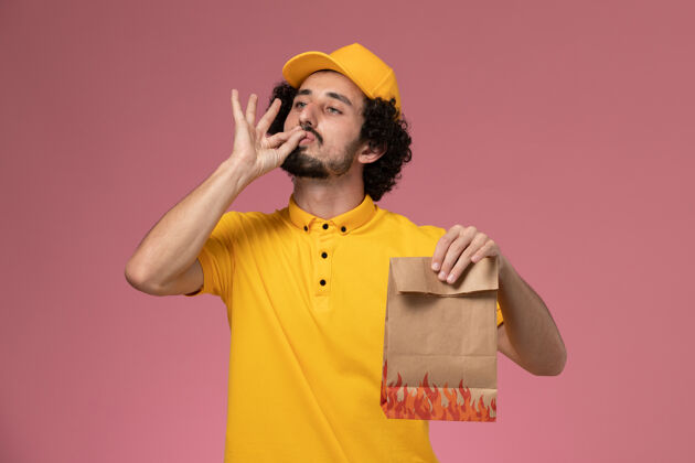 制服正面图：身穿黄色制服的男快递员手持纸质食品包装 在浅粉色墙上显示美味标志黄色持有送货