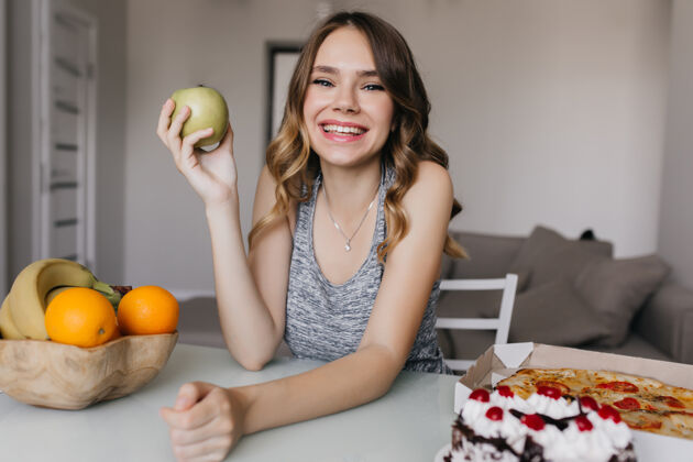 可爱欣喜若狂的年轻女士在早餐时吃着绿色的苹果和橘子积极的白人女孩吃水果和蛋糕的室内照片晚餐乐趣人