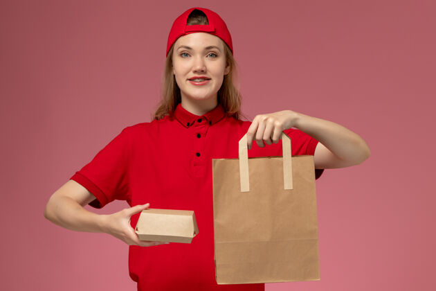快乐正面图：身穿红色制服 披风的年轻女快递员在粉色墙上拿着快递食品包成人微笑汉堡