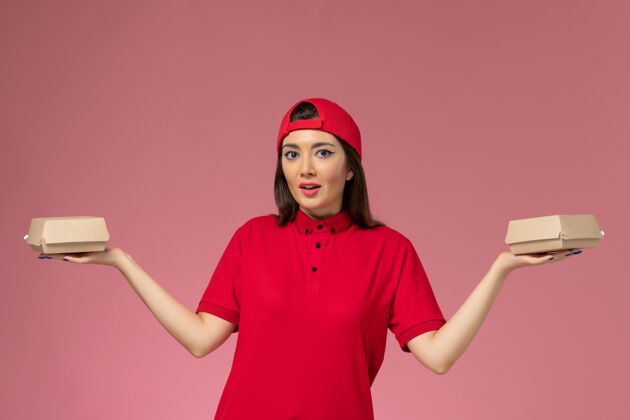 视图正面图：身穿红色制服和斗篷的年轻女快递员 手上拿着小包裹 放在粉红色的墙上人快乐手