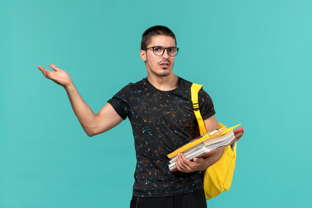 黑暗身穿深色t恤黄色背包的男学生正面图 浅蓝色墙上有文件和书籍档案人背包