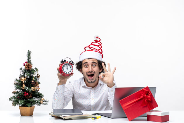 关心年轻的商人戴着圣诞老人的帽子 拿着钟 在办公室里展示了三个白色的背景礼物微笑圣诞节