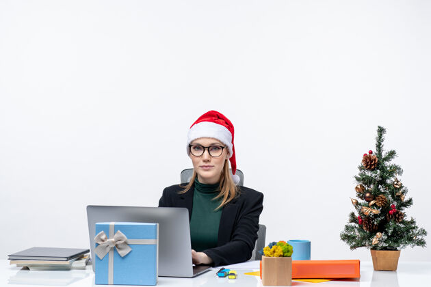 桌子骄傲快乐的女商人 戴着圣诞老人帽 坐在一张桌子旁 桌上放着圣诞树和礼物 在白色背景下查看邮件圣诞老人圣诞老人办公室