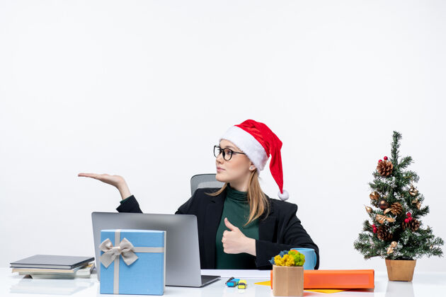 好的戴着圣诞老人帽的金发女商人坐在一张桌子旁 桌上放着圣诞树和礼物 做着“ok”手势 在白色背景下指着办公室右侧的东西右边圣诞树女人