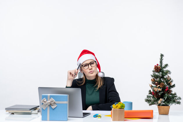 笔记本带着圣诞老人帽子的金发女人坐在桌子旁 桌上放着一棵圣诞树和一份白色背景的礼物圣诞老人电脑圣诞树