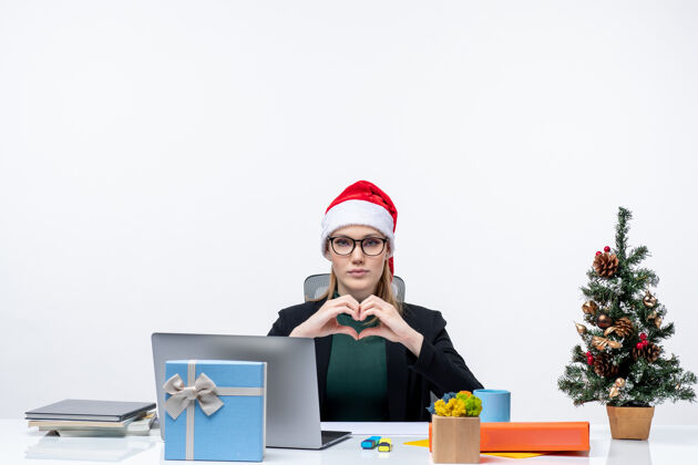女人带着圣诞老人帽子的浪漫女人坐在桌子旁 桌上放着一棵圣诞树和一份白色背景的礼物圣诞节帽子圣诞老人