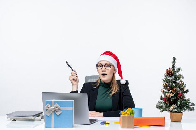 坐着一个好奇的金发女人 戴着圣诞老人的帽子 坐在一张桌子旁 桌子上放着一棵圣诞树和一份礼物 她把注意力集中在白色背景的东西上公司金发女郎帽子