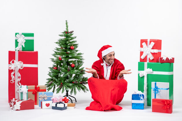 礼物有趣的年轻人打扮成圣诞老人与礼物和装饰圣诞树坐在地上的白色背景盒子年轻人年轻人