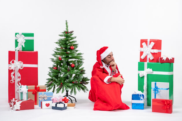 圣诞树体贴的年轻人打扮成圣诞老人 拿着礼物和装饰好的圣诞树坐在白色背景的地上年轻人圣诞圣诞老人