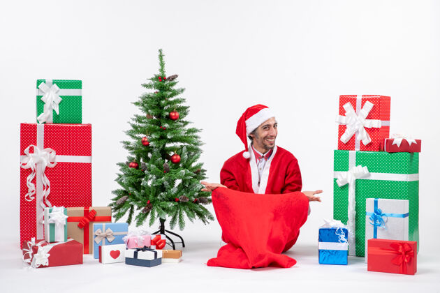 成人满意的年轻人打扮成圣诞老人与礼物和装饰圣诞树坐在地上的白色背景装饰年轻人微笑