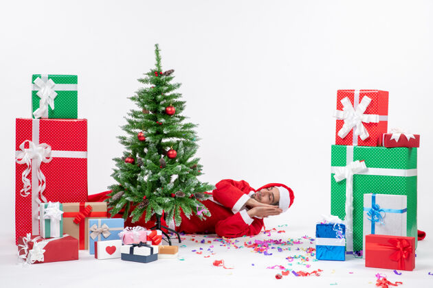 圣诞树喜庆的节日气氛与年轻的圣诞老人躺在圣诞树旁的礼物在不同颜色的白色背景股票照片冬青圣诞老人礼物