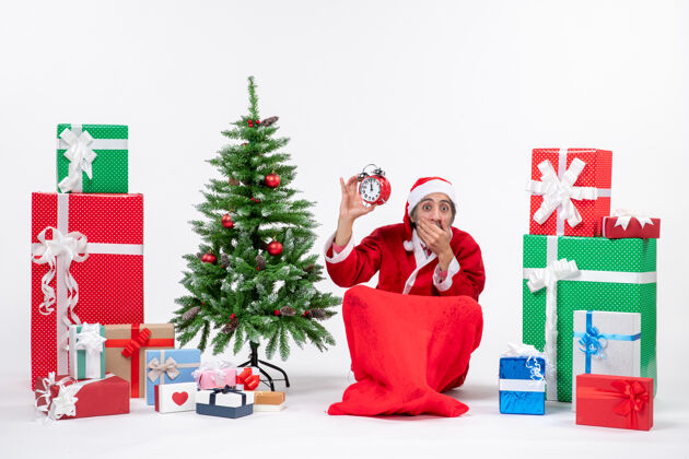 圣诞树吓坏了的圣诞老人坐在地上 在礼物和白色背景装饰圣诞树旁展示着时钟圣诞老人害怕礼物