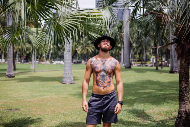微笑欧洲帅哥纹身 大胡子 戴着经典帽子 赤裸上身 在热带公园的赛场上健康自然男子汉