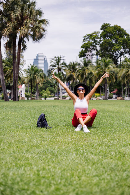 时尚快乐女人背着背包环游曼谷 在绿草如茵的热带公园里享受美丽的晴天树热带度假