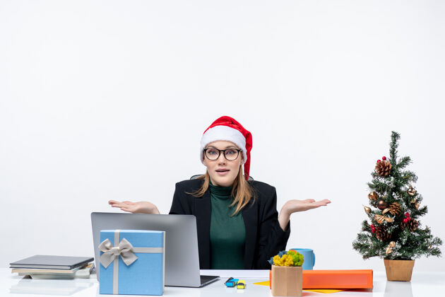 圣诞老人带着圣诞老人帽子的积极好奇的女商人坐在一张桌子旁 桌子上有一棵圣诞树和一份白色背景的礼物电脑商务帽子