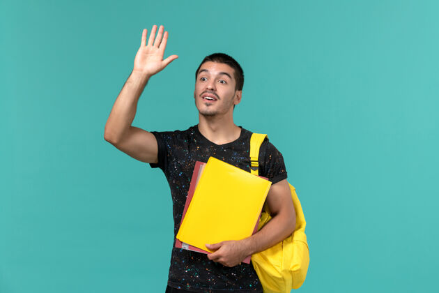 学生身穿深色t恤黄色背包的男学生的正面图 在淡蓝色的墙上挥舞着不同的文件微笑背包人