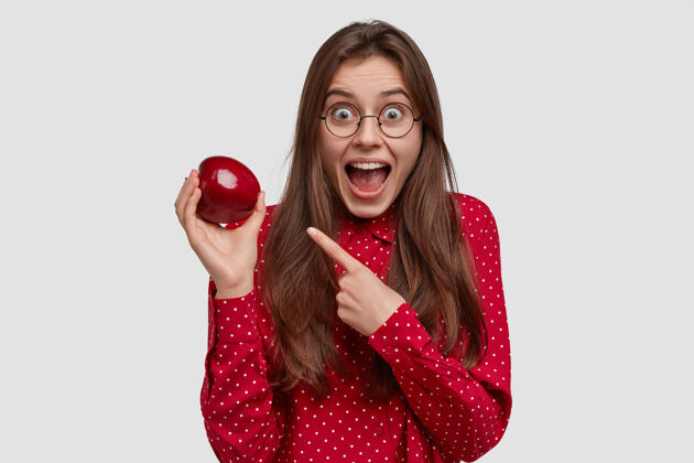 美味快乐的年轻女士指着鲜红多汁的苹果 展示健康的食物 坚持节食 下巴下垂 留着长发 惊讶的表情外表指着节食