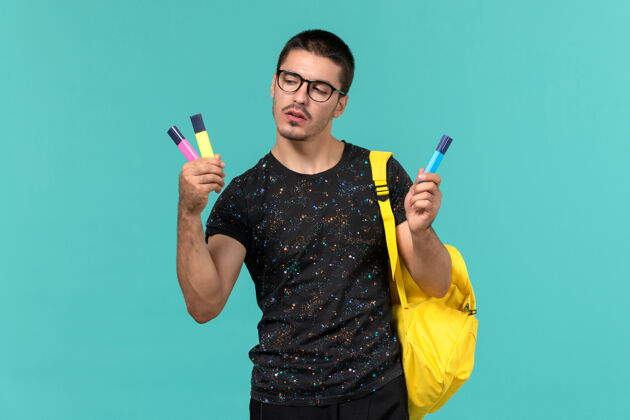 甜点身穿深色t恤 黄色背包 蓝色墙上拿着彩色毛毡笔的男生正面图钢笔毡男学生