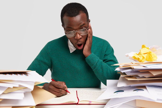 工作场所惊艳的黑人男性横观用文件工作 在记事本上记笔记 有惊呆的表情 戴着大眼镜工作Omg报告