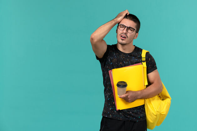 背包前视图中的男学生穿着深色t恤黄色背包拿着不同的文件和咖啡思考蓝色墙上持有学生大学