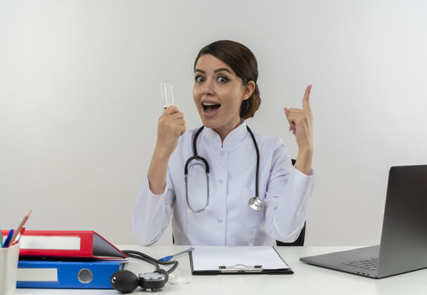 工作快乐的年轻女医生穿着医用长袍 带听诊器 坐在办公桌旁 拿着医疗工具 拿着灯泡 大拇指竖立在有复印空间的白色隔离墙上灯光女电脑