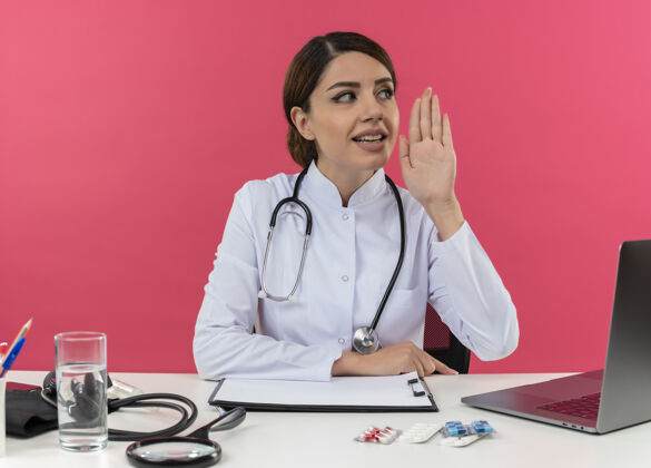 医疗高兴的年轻女医生穿着医用长袍 听诊器坐在办公桌上 用电脑和医疗工具打电话给有复印空间的人长袍穿着电脑