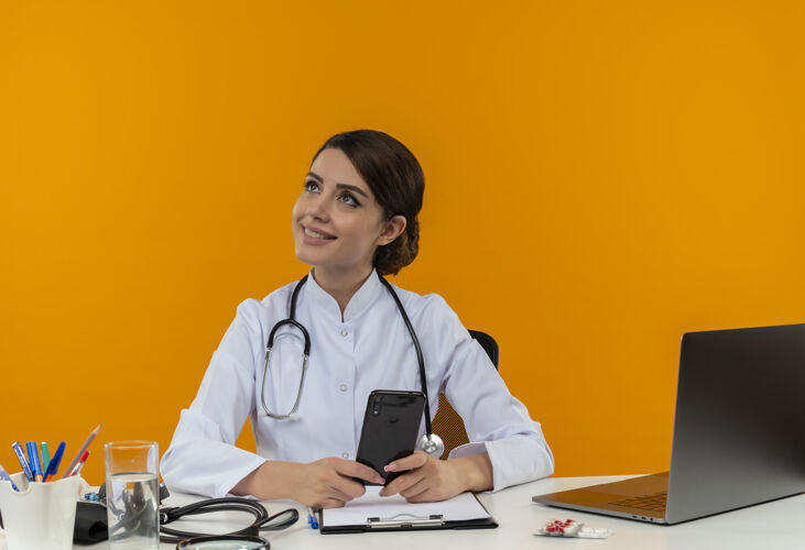 电脑看着面带微笑的年轻女医生 穿着医用长袍 手持听诊器 坐在办公桌旁 拿着医疗工具 拿着电话 站在隔离的黄色墙上穿着年轻人隔离