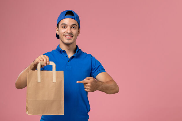 专业正面图：身穿蓝色制服和披风的年轻男性快递员 手上拿着一个纸包裹 放在粉红色的墙上人粉色包裹