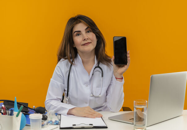 办公桌高兴的中年女医生穿着医用长袍 带听诊器 坐在办公桌旁 用笔记本电脑和医疗工具 把电话放在隔离的橙色墙上 还有复印空间工作工具抱着