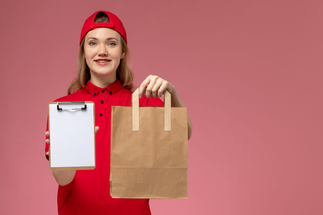 服务正面图：身穿红色制服的年轻女快递员拿着快递食品包 在浅粉色的墙上放着记事本人女微笑